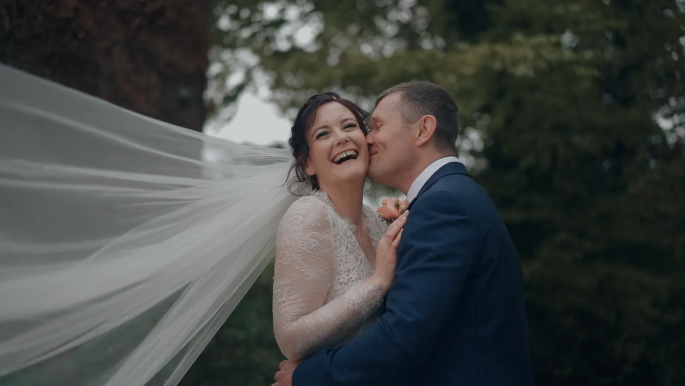 A joyful bride in a white lace gown with a flowing veil and groom in a navy suit embrace and kiss outdoors under green trees, with the veil dramatically billowing in the wind.
