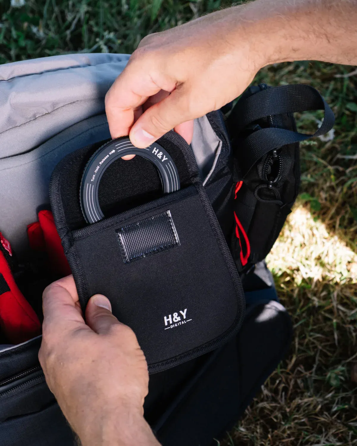 A photographer’s hands placing an H&Y Revoring adapter back into its branded soft case inside an open camera bag, packed with other gear and resting on grass outdoors.