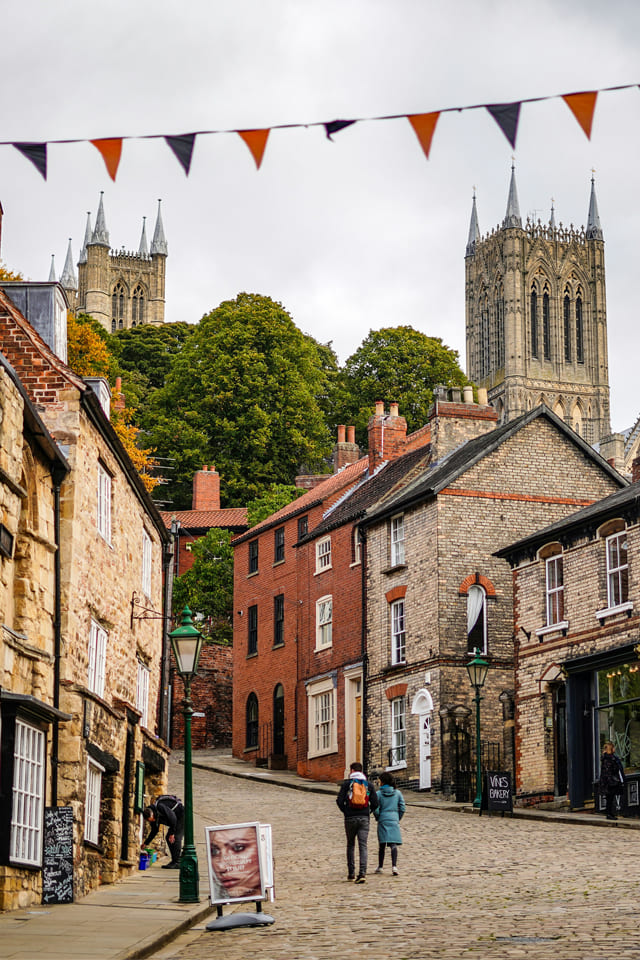 Steep cobbled street in Lincoln lined with old brick and stone houses leading up to Lincoln Cathedral, with bunting overhead and people walking.