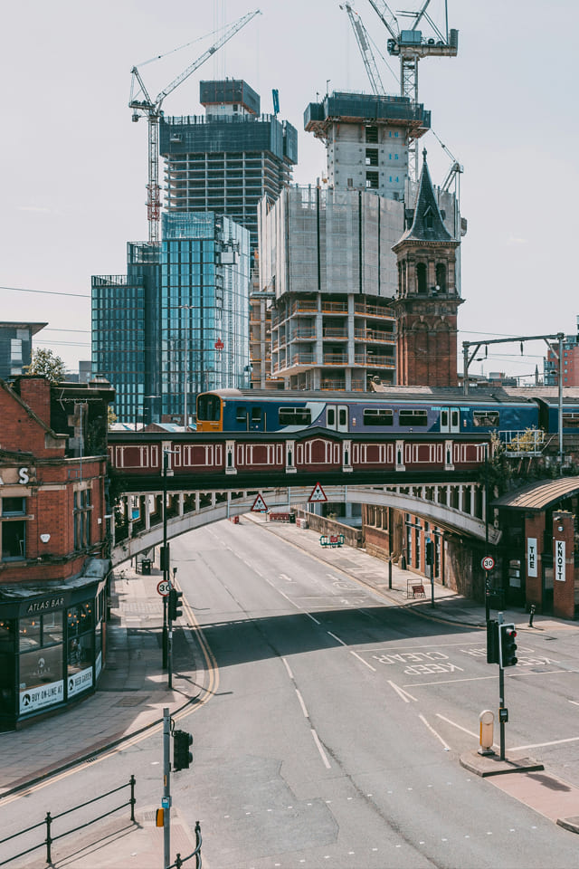 Urban scene of a train crossing an elevated red bridge in front of modern high‑rise construction cranes and older brick buildings on a wide city Manchester street.